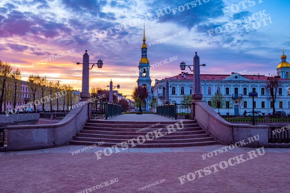 Фотография запечатлела вечерний Санкт-Петербург с видом на Никольский морской собор, окрашенный в мягкие тона заходящего солнца. Живописное небо с переходами от розового к голубому создает почти магическую атмосферу. Сцена идеально подходит для публикаций о городе, в туристических материалах или как элемент художественной фотографии для интерьера.